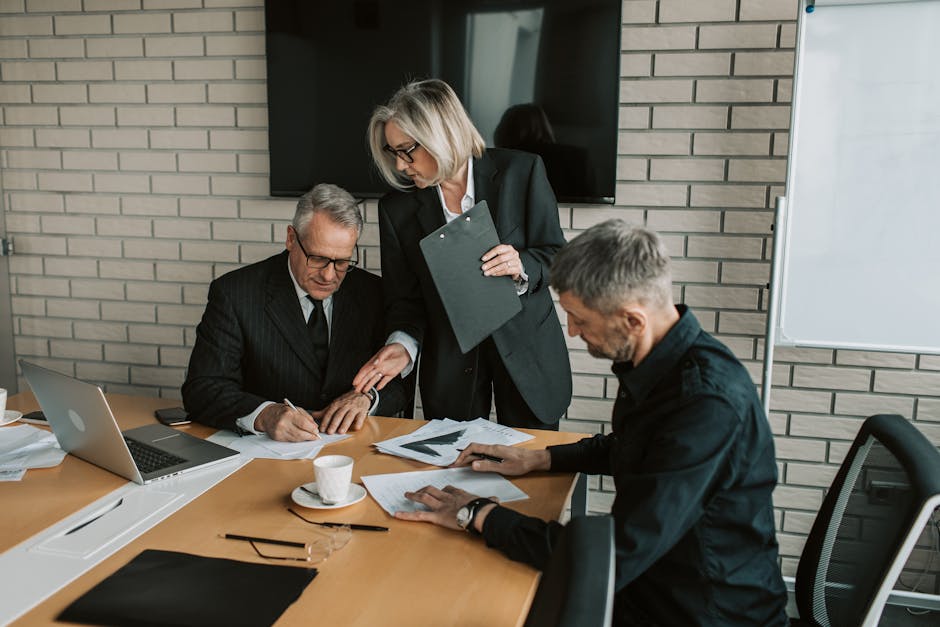 Three mature professionals in a business meeting discussing and signing documents in an office setting.